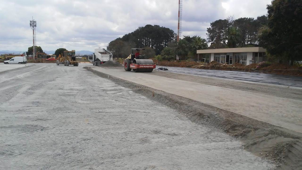 Rolo compactador na base do pavimento, com motoniveladora e caminhão ao fundo